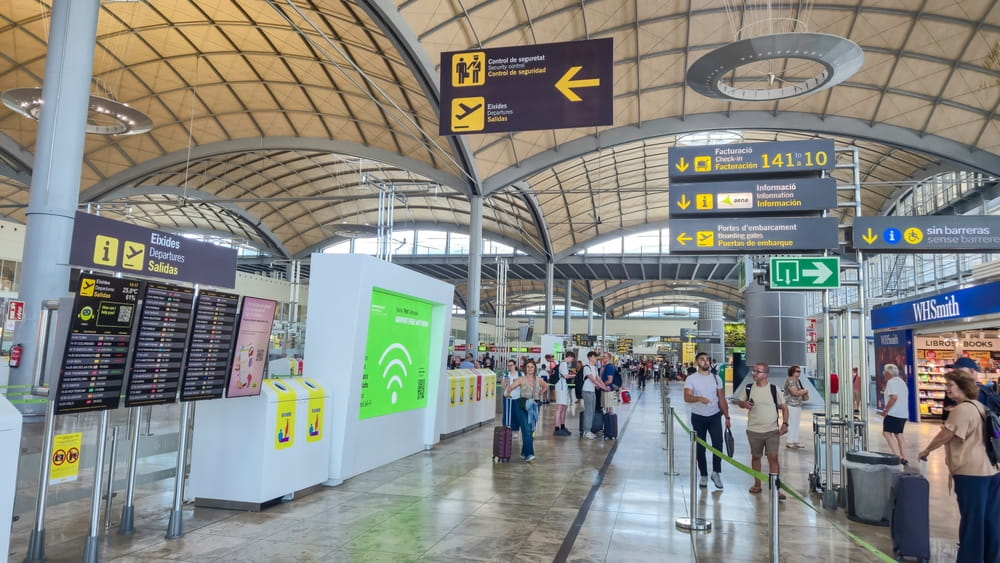 Interior of Alicante Airport departures hall with security, check-in signs, flight information boards and passengers"