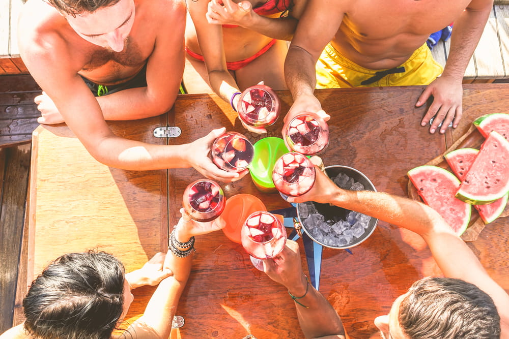 Amigos brindando con copas de sangría roja en España en una mesa soleada de verano con sandía y hielo