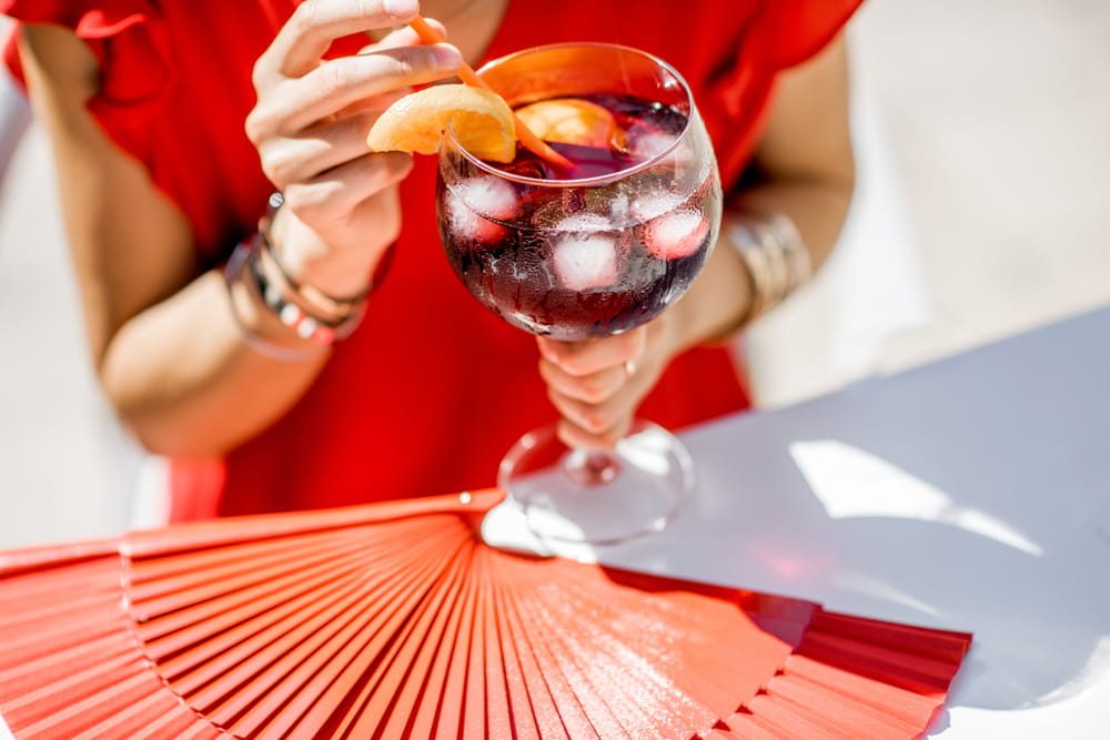 Mujer sosteniendo una copa fría de sangría roja con rodajas de naranja y hielo junto a un abanico rojo